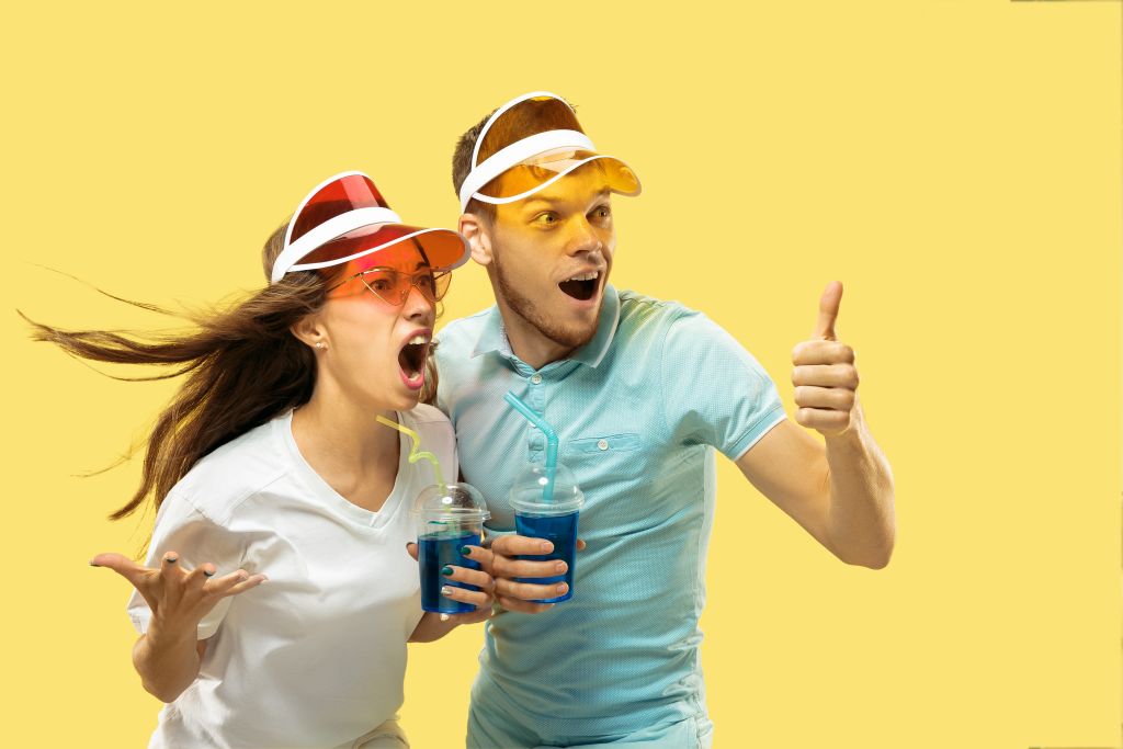 Beautiful young couple's half-length portrait isolated on yellow studio background. Woman and man standing with drinks in colorful caps. Facial expression, summer, weekend concept. Trendy colors.